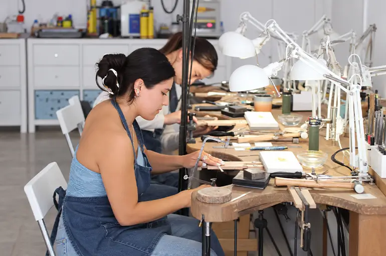 Two jewelers working at their benches in a jewelry workshop, crafting custom jewelry pieces by hand.